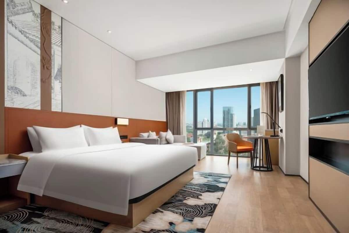 A modern hotel room with a large bed, a desk, a chair, a TV, and a view of the city.