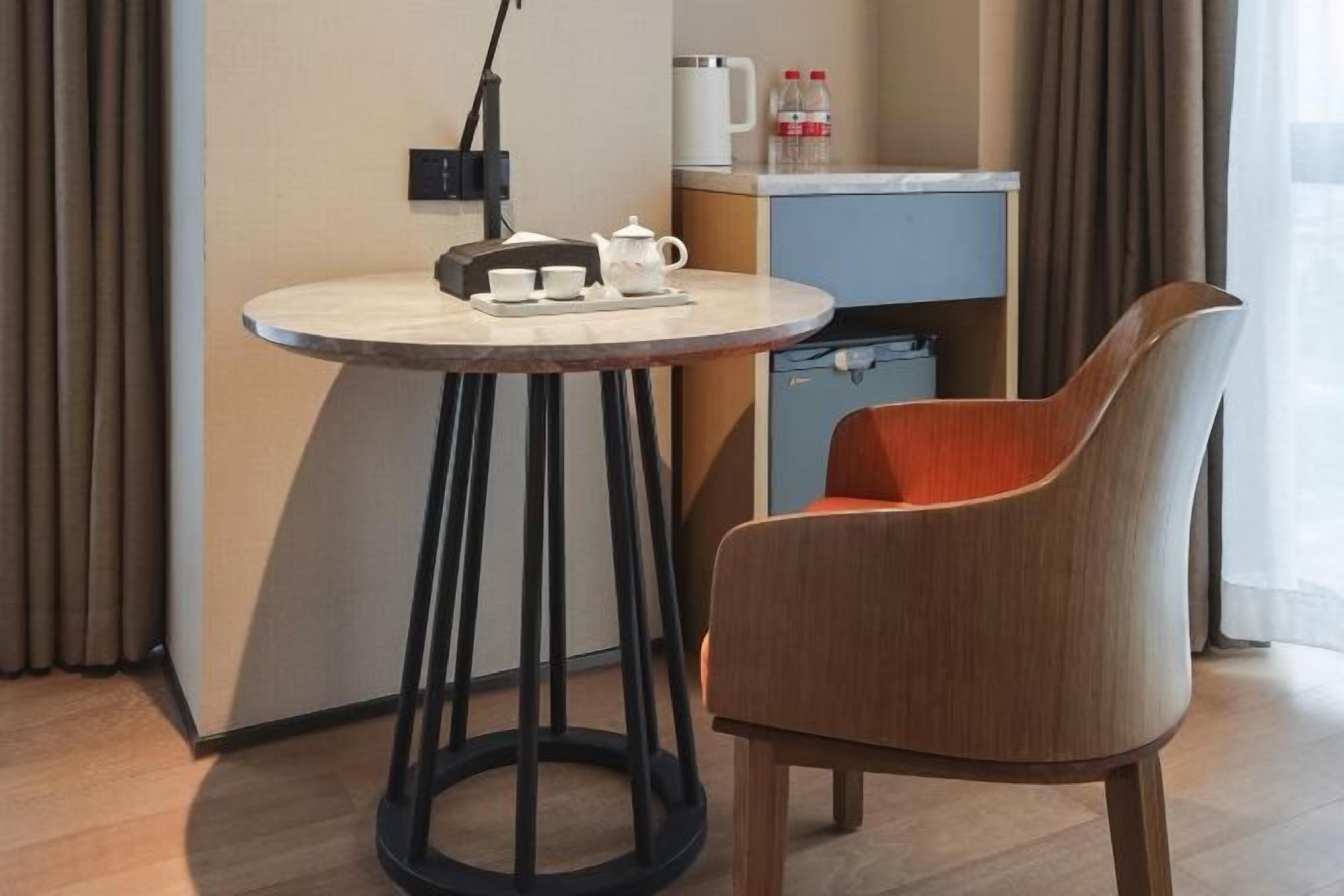 Een kleine ronde tafel met een theepot, kopjes en een dienblad, een moderne stoel, een kitchenette en een raam met gordijnen.