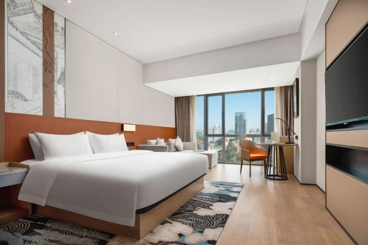 A modern hotel room with a large bed, a desk, a chair, a TV, and a city view.