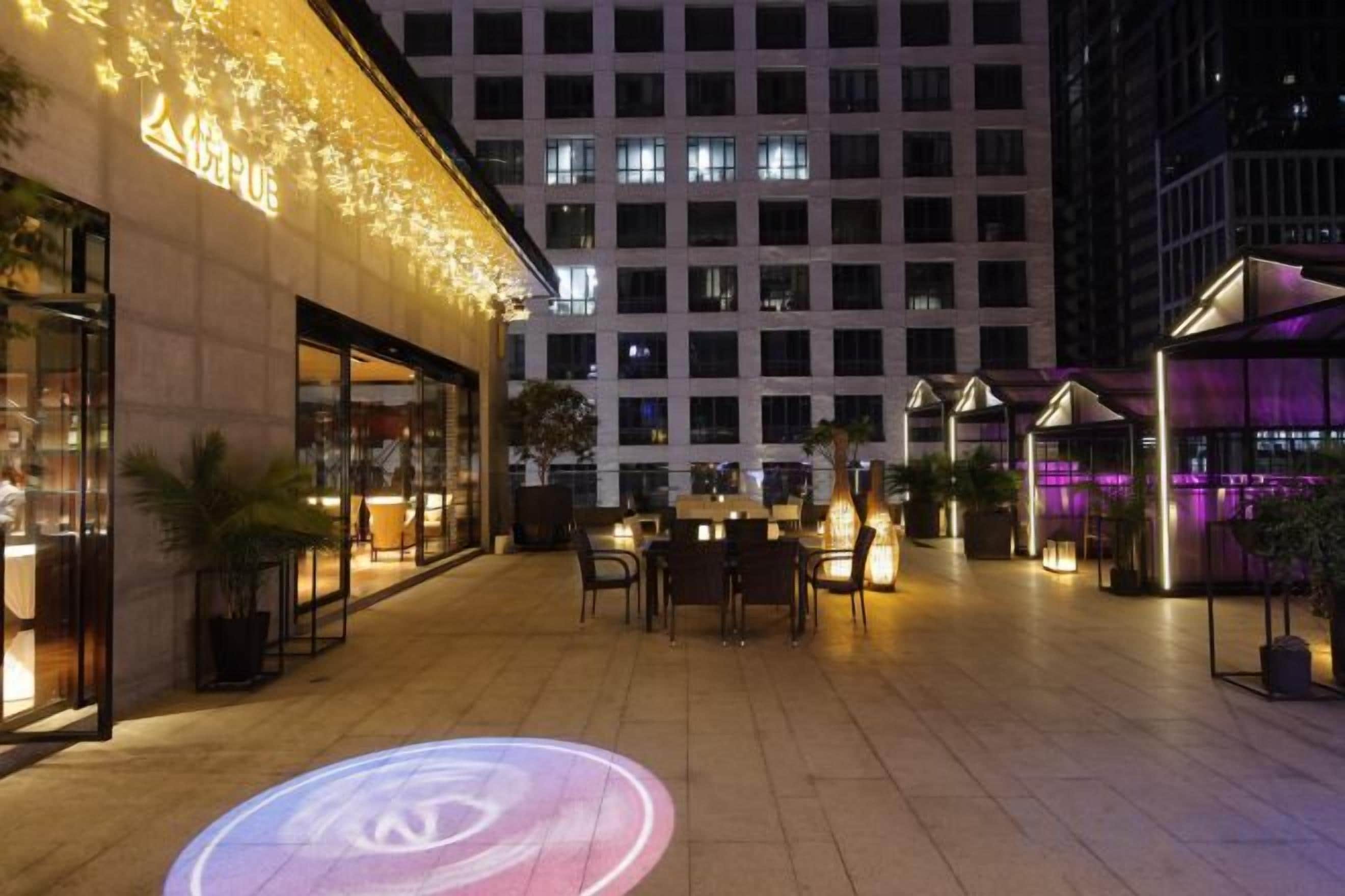 An outdoor urban setting with a building facade, outdoor seating area, and a circular light projection on the ground.
