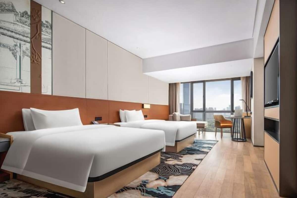 A hotel room with two beds, a desk, a chair, a TV, and a large window offering a city view.