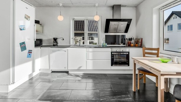 A modern kitchen with white cabinets, a built-in oven, a microwave, and a coffee machine. There is a dining table with chairs and a window providing a view of the outside. - "Ullakarin" - 200m from the sea by Interhome (Thyborøn)