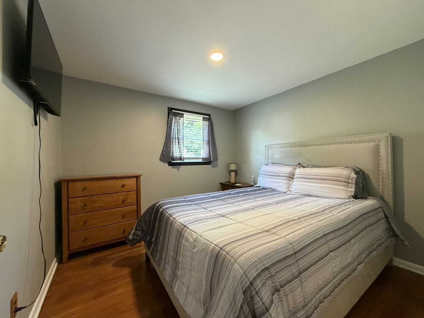 Een slaapkamer met een bed, een houten dressoir, een nachtkastje, een lamp en een raam met gordijnen.
