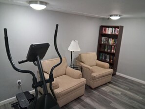 A room with a treadmill, two armchairs, a small table with a lamp, and a bookshelf.