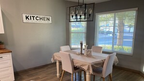 A dining area with a table set for four, a kitchen sign on the wall, and a view of the outdoors through large windows. - Modern Glendale Getaway /Mins from I-17 & 101 / 6 Flags, Hiking & Golf close by (Glendale)