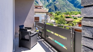 Un balcón con vistas a un paisaje montañoso, una silla de mimbre y una mesa con una botella y copas.