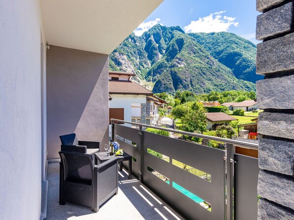 A balcony with a view of a mountainous landscape, a wicker chair, and a table with a bottle and glasses.