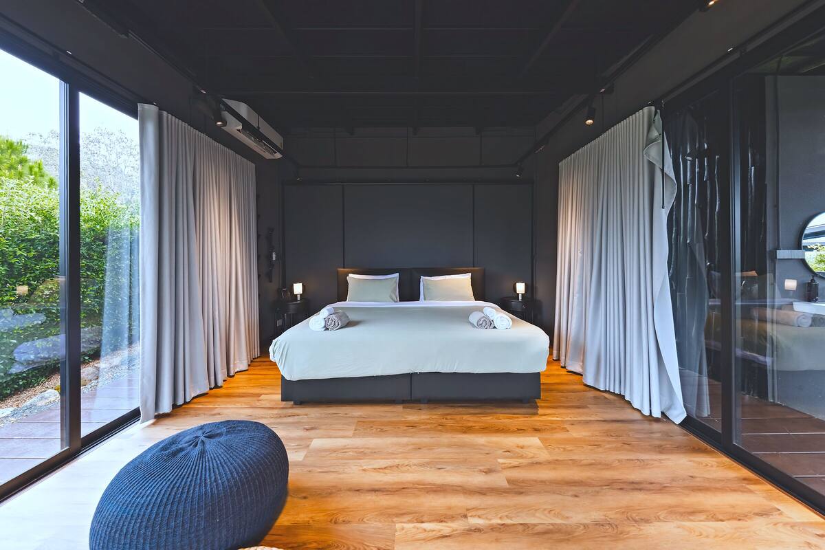 A modern bedroom with a large bed, bedside lamps, and a view through floor-to-ceiling windows.