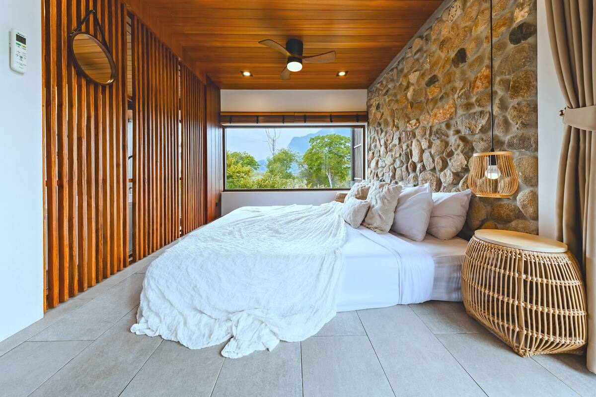 A bedroom with a large bed, wooden slats, a stone wall, and a view of trees and mountains.