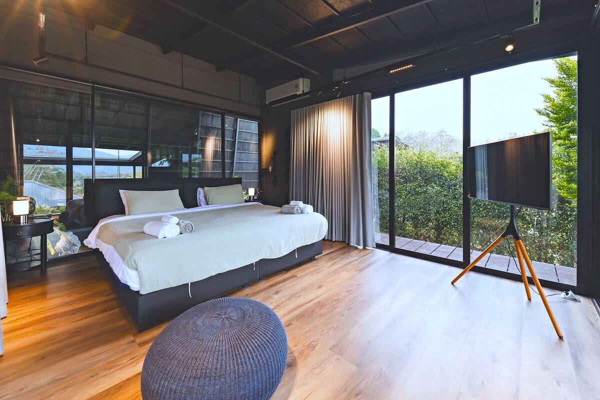 A modern bedroom with a large bed, a flat-screen TV, and a view of greenery through large windows.