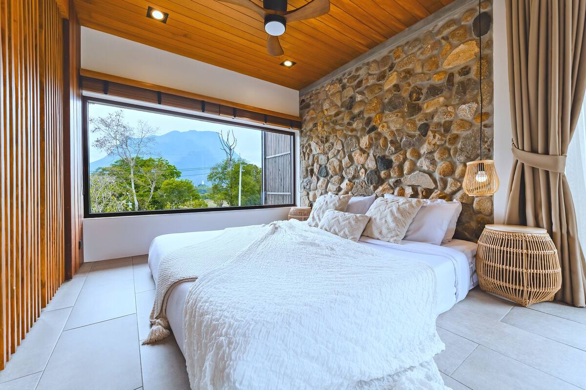 A bedroom with a large bed, a stone wall, a wooden ceiling, and a view of trees and mountains.