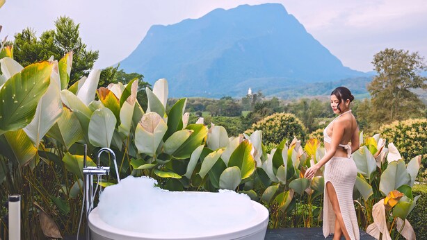 Una mujer de pie junto a una bañera grande en un jardín, con una montaña al fondo.