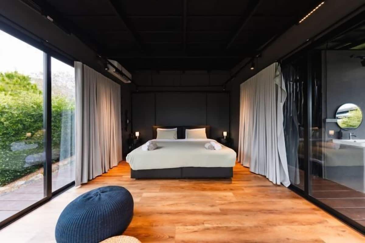 A modern bedroom with a large bed, a seating area, and a view of greenery outside.