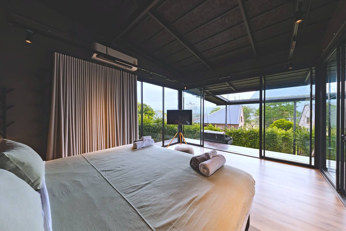 A modern hotel room with a large bed, a TV, and a view of greenery and a building outside.