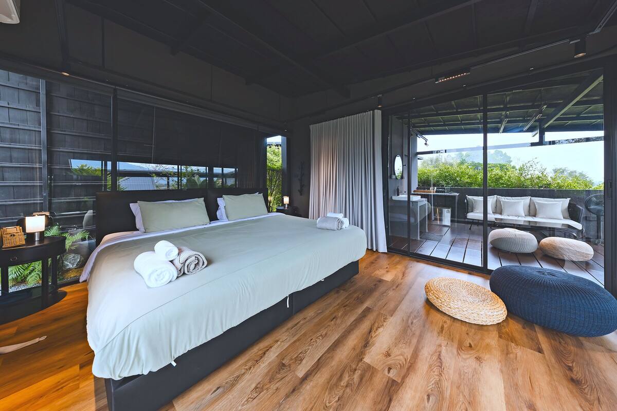 A modern bedroom with a large bed, a glass door leading to a balcony with seating, and a view of greenery.