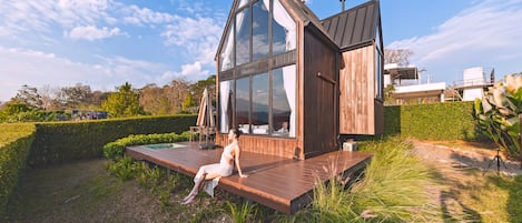 Una casa moderna con fachada de vidrio, terraza de madera y un pequeño espacio de estar al aire libre.