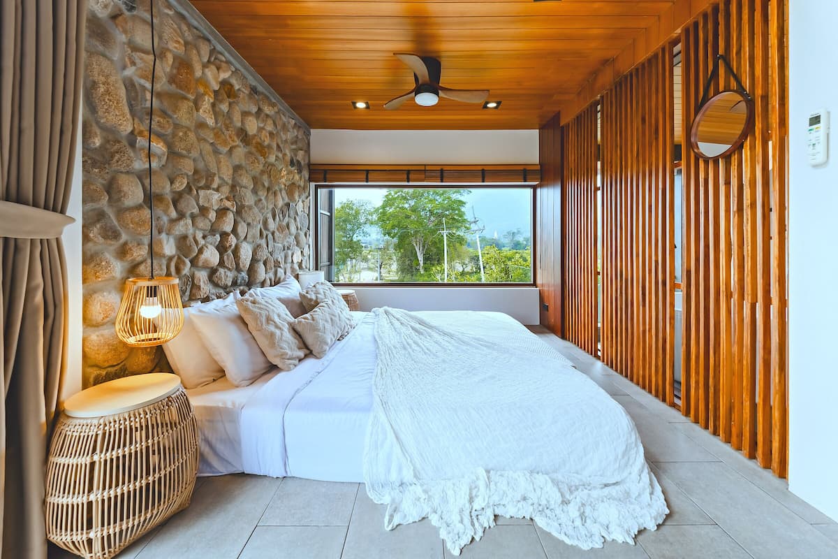 A bedroom with a stone wall, wooden ceiling, a bed with white bedding, a woven stool, and a view of greenery outside.