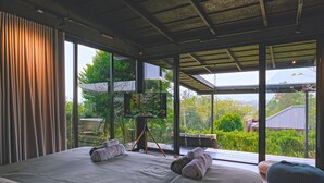Un dormitorio moderno con una cama grande, un televisor sobre un trípode y vistas a zonas verdes al exterior.