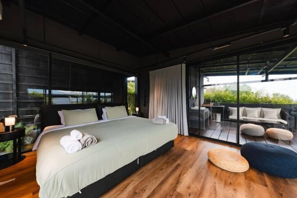 A modern hotel room with a large bed, a balcony with seating, and a view of greenery.
