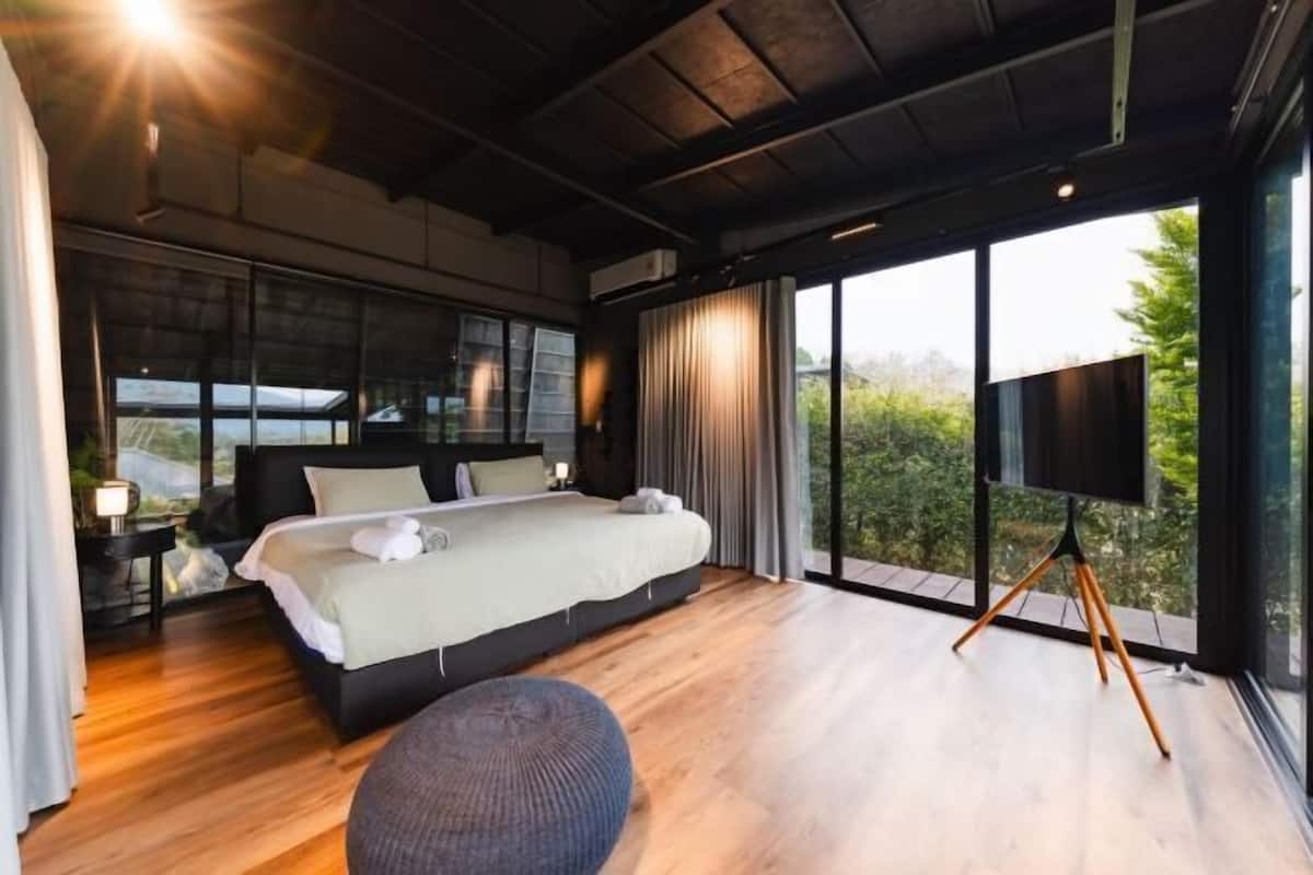 A modern bedroom with a large bed, a TV on a tripod stand, and a view of greenery through large windows.