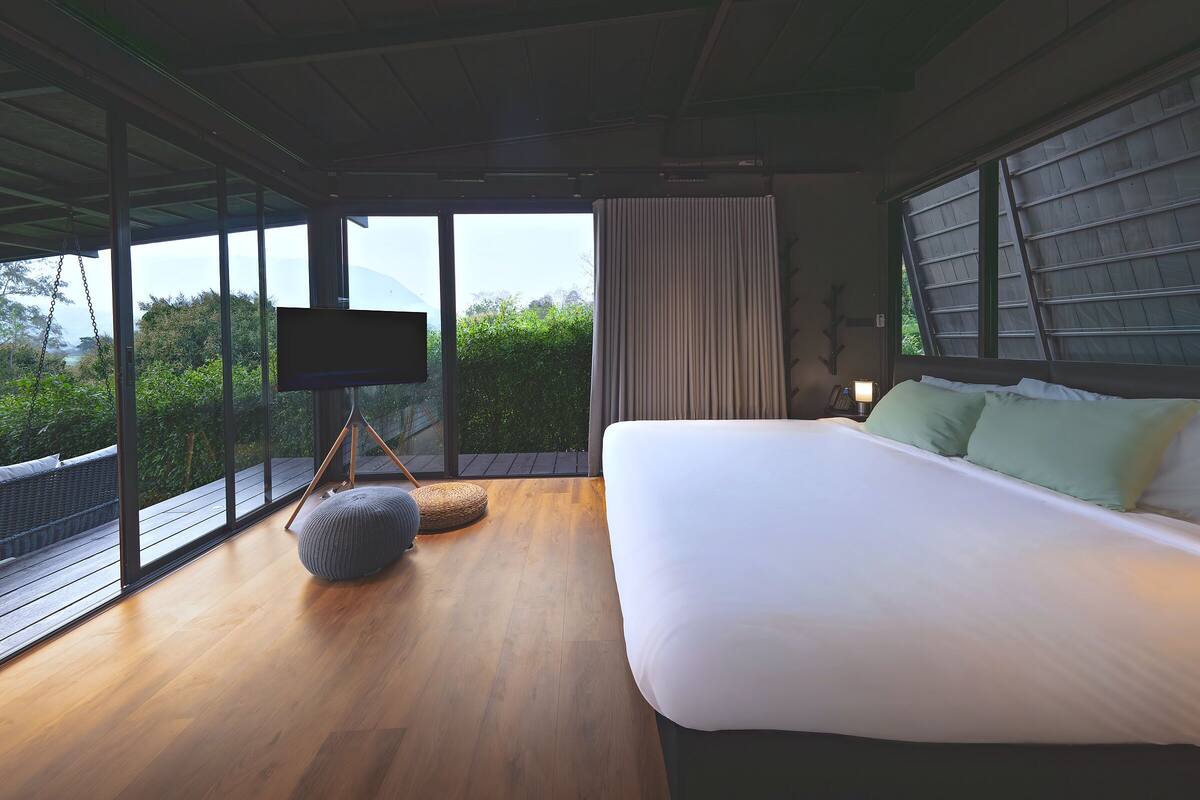 A bedroom with a large bed, a television on a stand, a bedside table with a lamp, and a view of greenery through large windows.