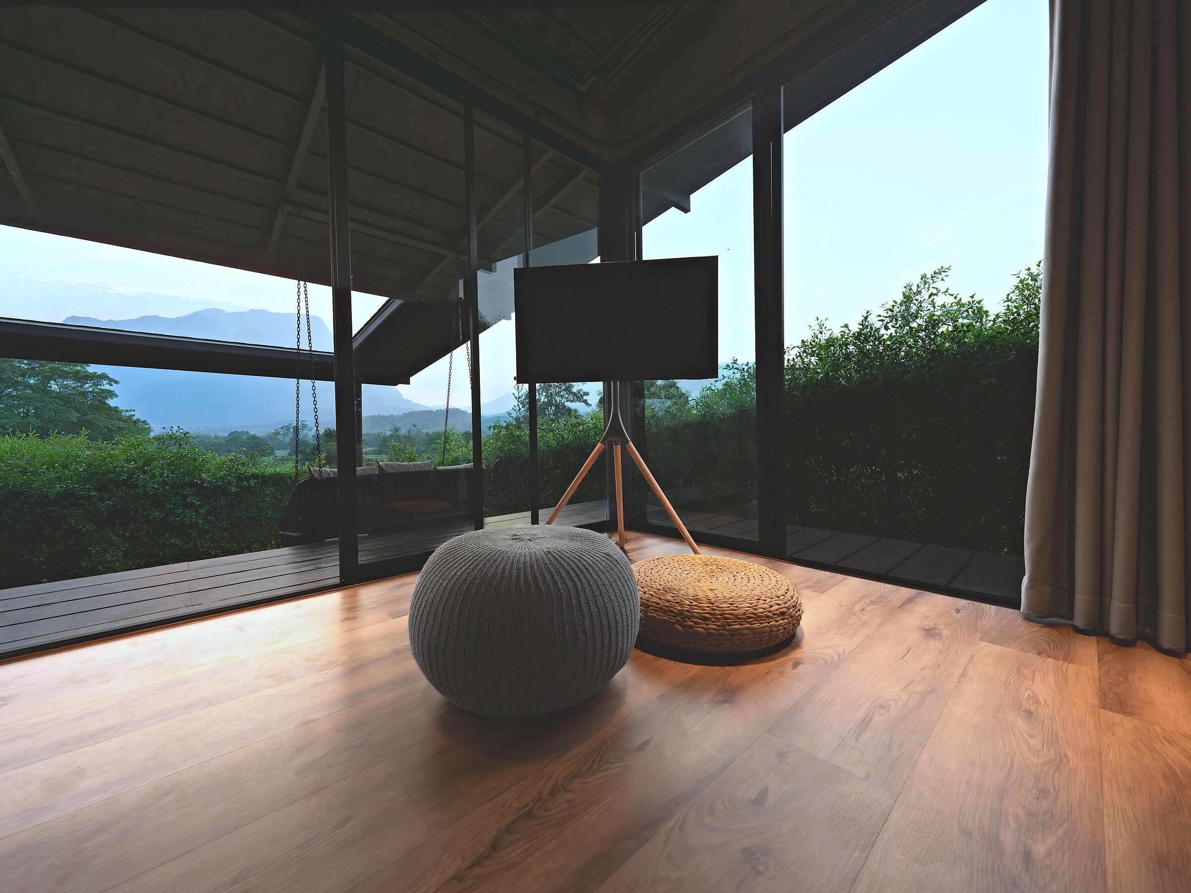 A modern room with a large window, wooden flooring, a grey knitted ottoman, a woven pouf, and a tripod-mounted screen.