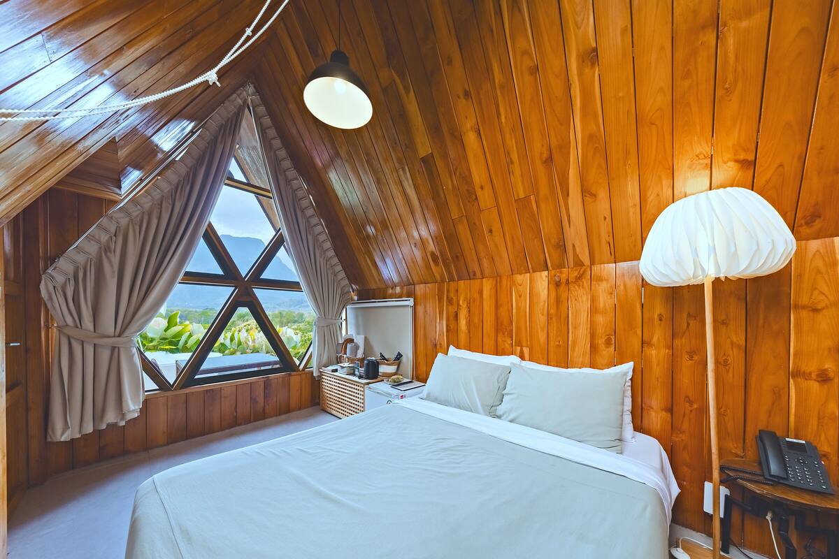 A bedroom with a large bed, wooden walls, a window with curtains, a bedside table, and a lamp.
