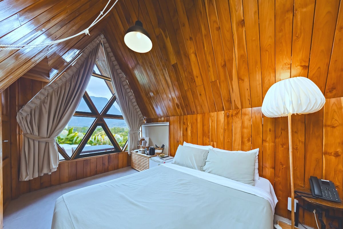 A bedroom with a large bed, wooden walls, a window with curtains, a bedside table, and a lamp.