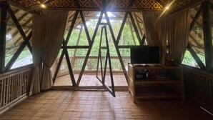 A room with a wooden floor, a TV on a wooden stand, and a view of greenery through large windows. - The Osing Bamboo Resort - Banyuwangi (Banyuwangi)