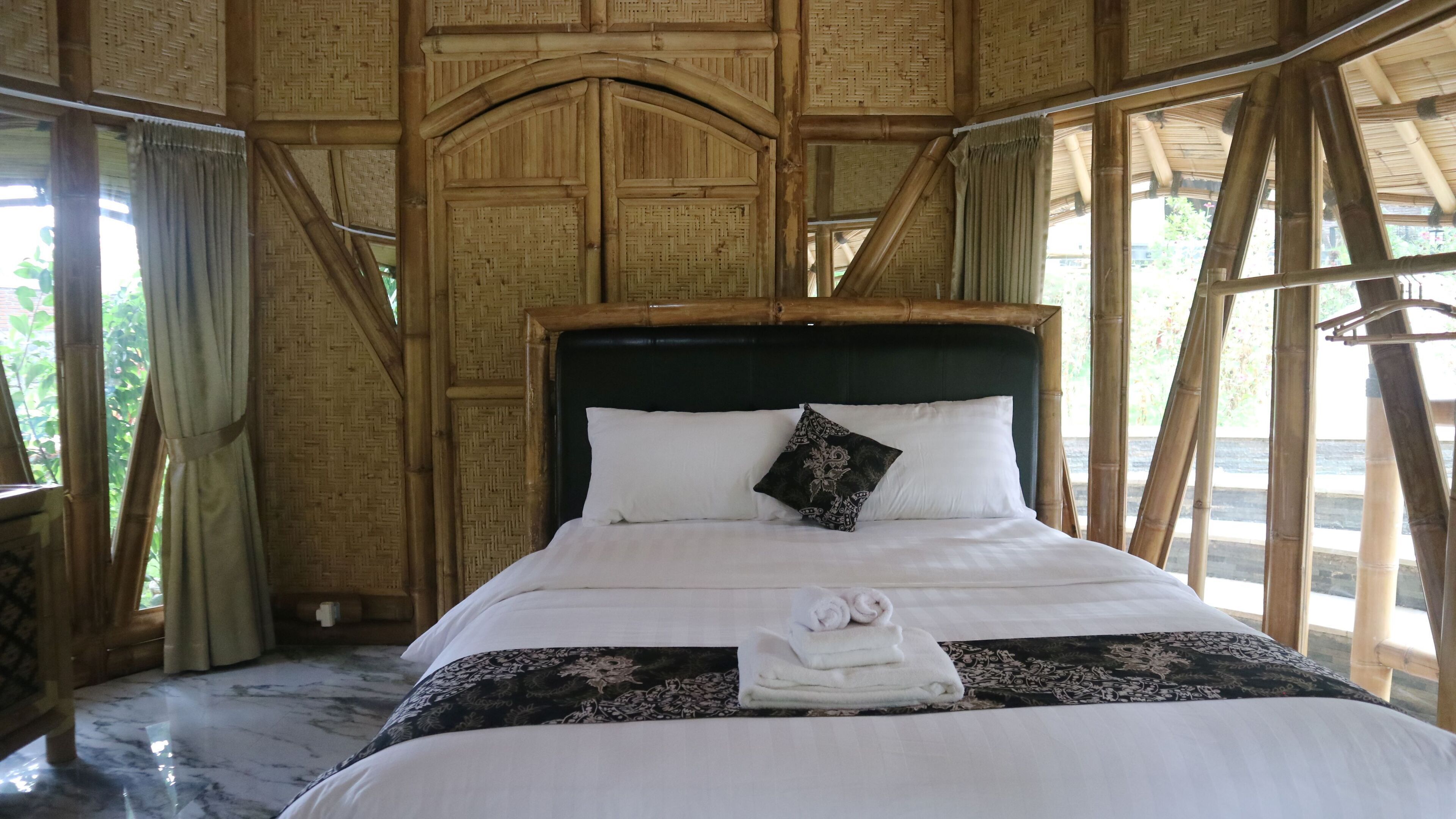 A bed with white linens and a black patterned pillow, a folded towel, and a dark headboard in a room with bamboo walls and a window.