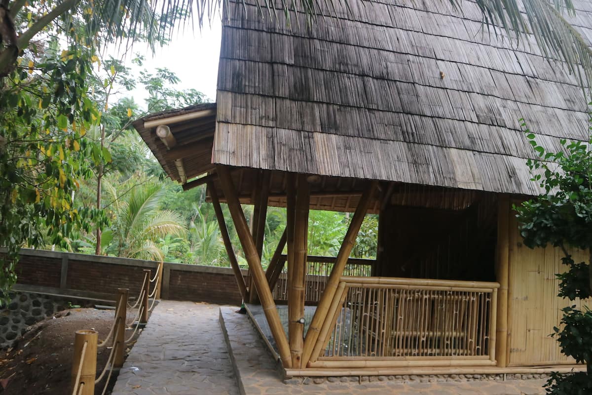 A rustic wooden structure with a thatched roof, bamboo railings, and a stone pathway leading to it.