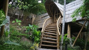 A spiral staircase made of bamboo leading to a thatched-roof structure surrounded by lush greenery.