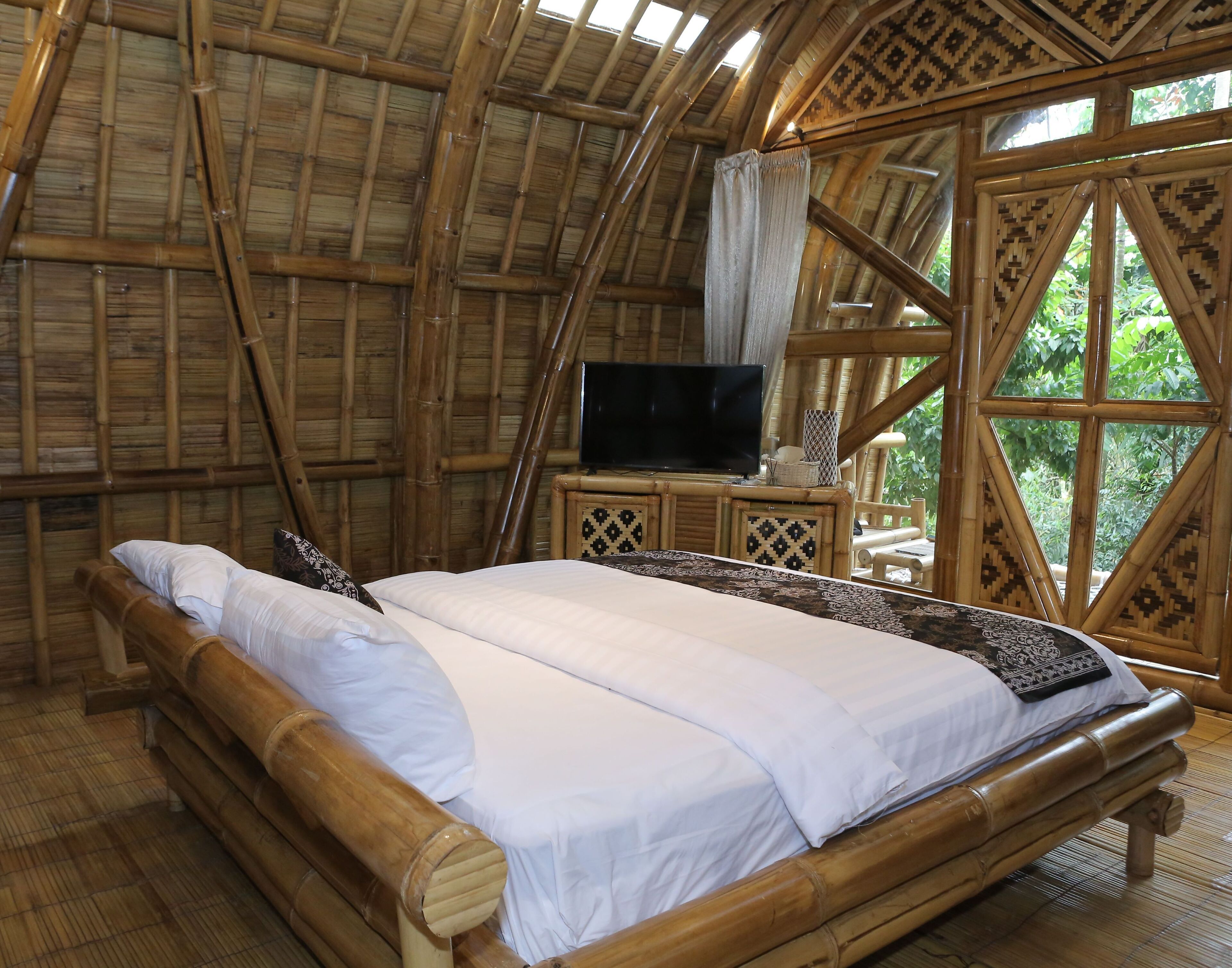 A bamboo-framed room with a bed, a television, and a window with a view of greenery.