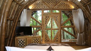 A room with a large bed, a television, and a window with a view of trees. - The Osing Bamboo Resort - Banyuwangi (Banyuwangi)