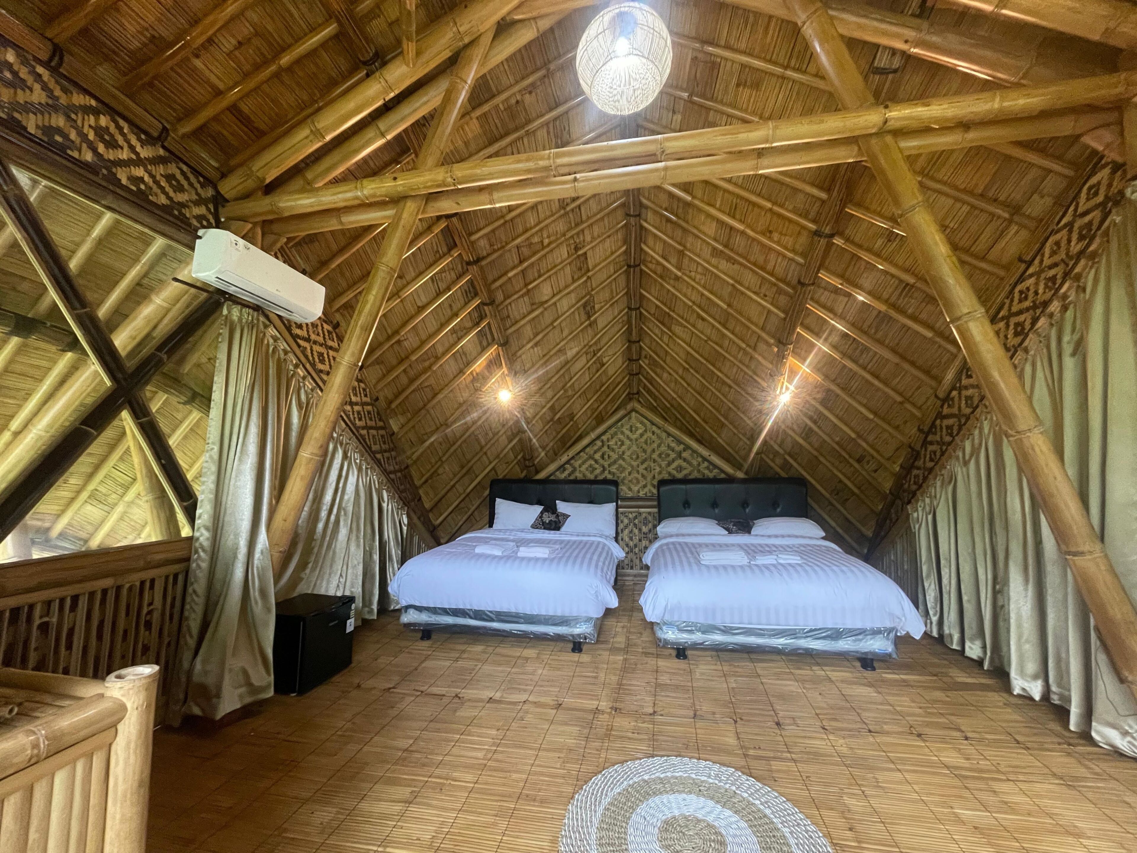 A room with two beds, a wooden floor, a high ceiling with bamboo beams, and a large circular light fixture.