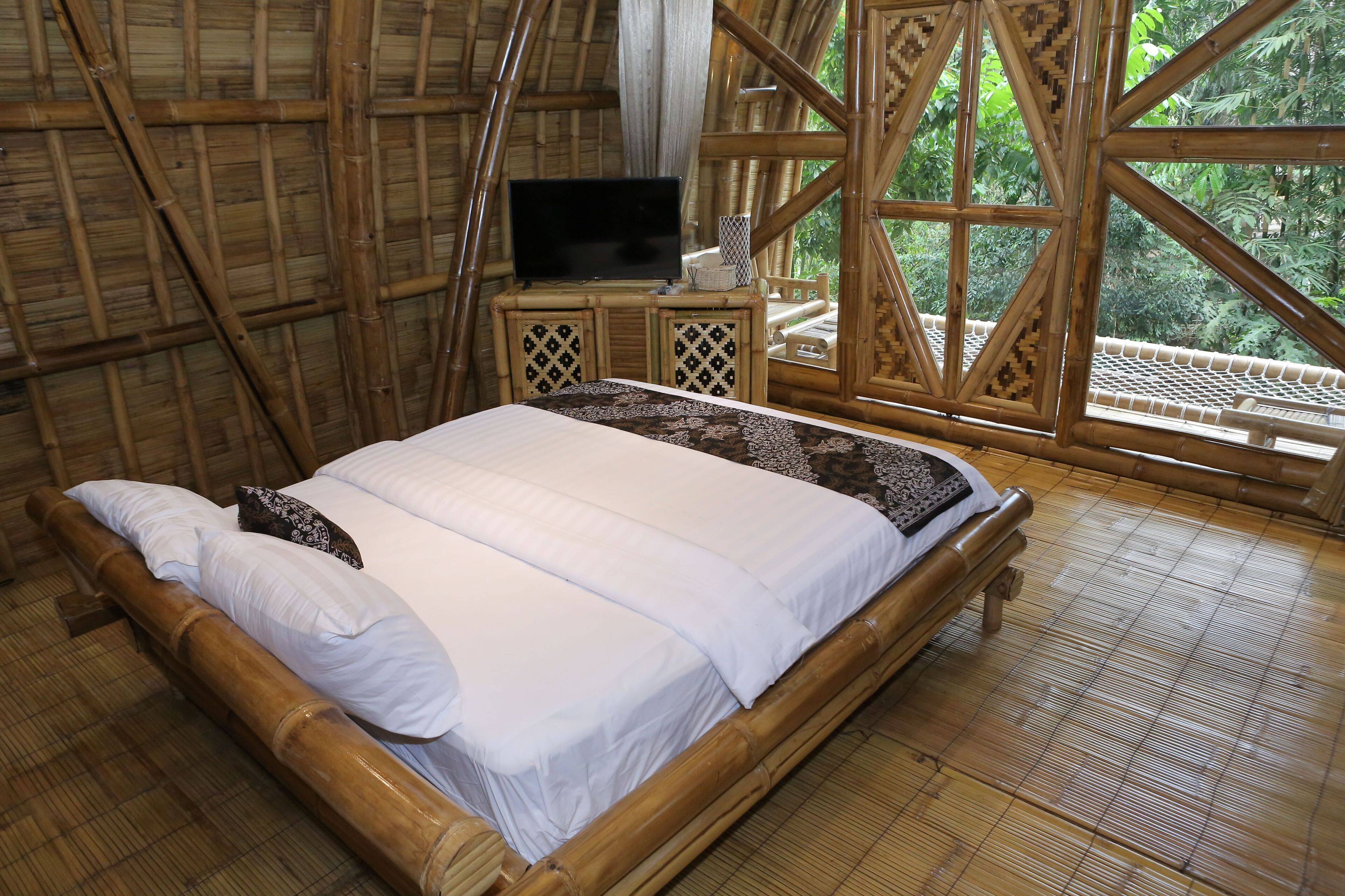 A room with a bed, a television on a wooden nightstand, and large windows with a view of greenery.