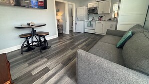 Uma sala de estar moderna com um sofá cinzento, uma pequena mesa de jantar com banquetas e uma cozinha com armários e eletrodomésticos brancos.