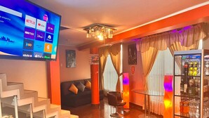 Un vestíbulo de hotel con una televisión de pantalla plana mostrando servicios de streaming, una escalera, un sofá negro, una mesa de vidrio y un puesto de bebidas.