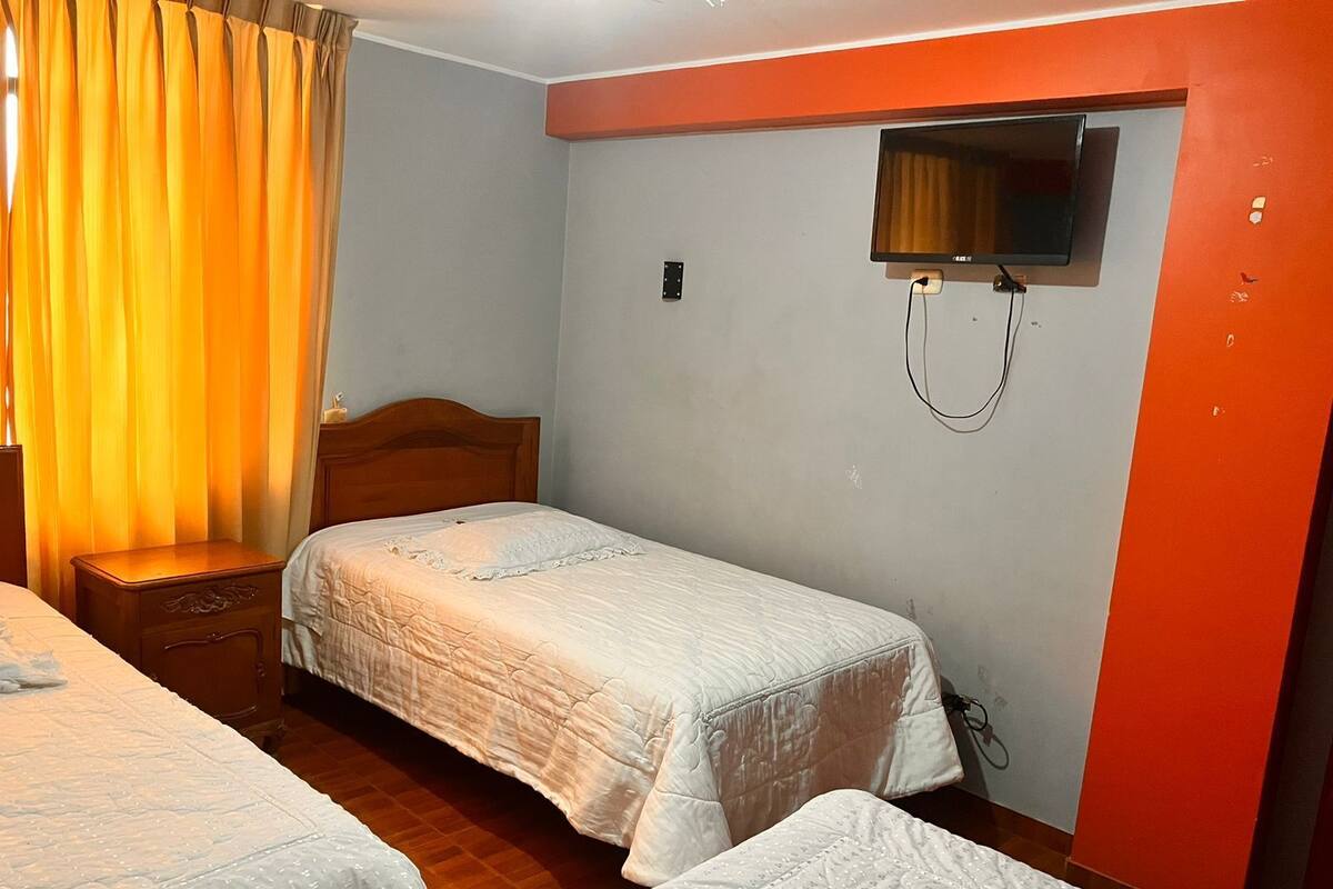 Une chambre avec deux lits, une table de chevet en bois, un téléviseur fixé au mur et un rideau à la fenêtre.