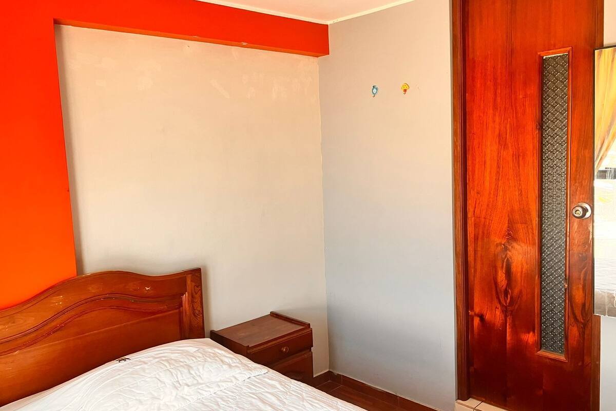 Une chambre avec un lit en bois, une table de chevet, une porte dotée d’un panneau texturé et un mur avec un détail rouge.