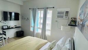 A hotel room with a bed, a desk with a TV, a window with curtains, an air conditioning unit, and a wall with a decorative piece. - Amourette's Studio at Mesaavirre Garden (Bacolod)