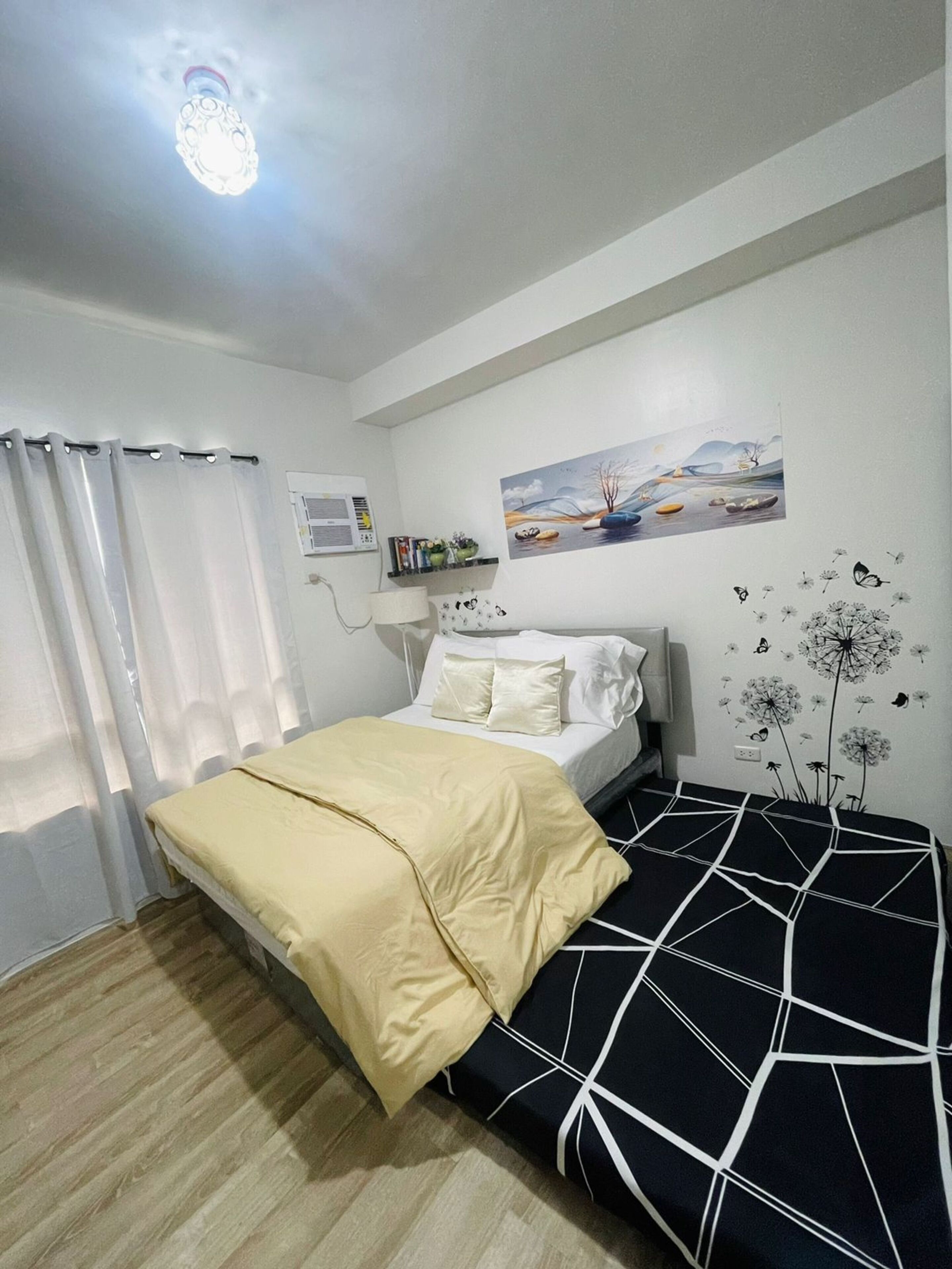 Un dormitorio con cama, mesita de noche, lámpara, ventana con cortinas, aire acondicionado instalado en la pared y un cuadro.