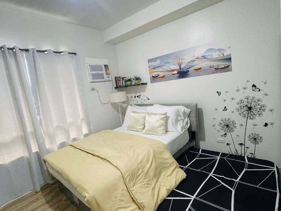 Un dormitorio con cama, mesita de noche, lámpara, ventana con cortinas, aire acondicionado instalado en la pared y un cuadro.