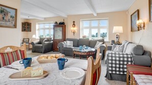 A cozy living room with a dining area, featuring a sofa set, a coffee table, and a dining table with a cheese platter and bread.