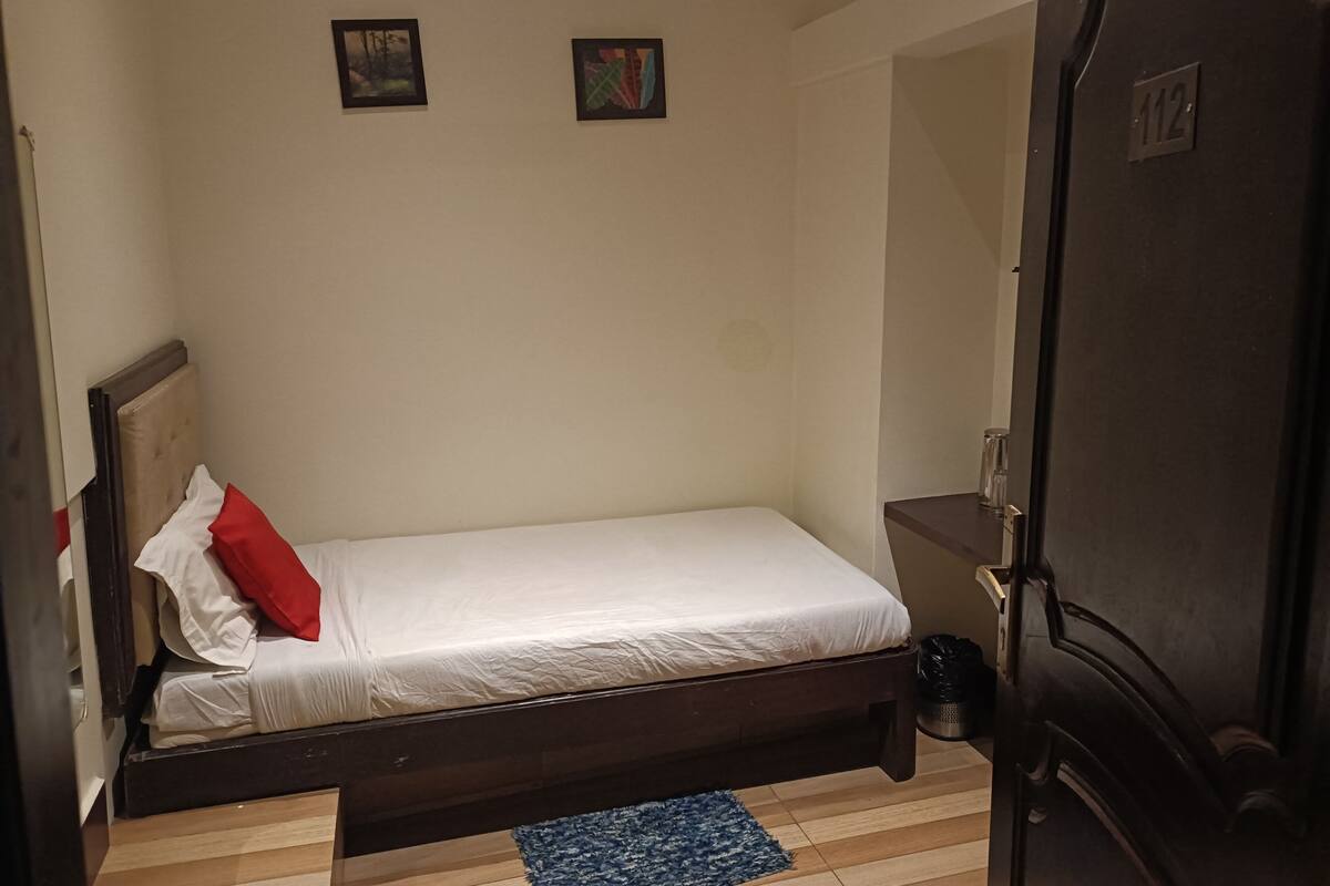A small, single-bed room with a wooden headboard, a white mattress, and a red pillow. There are two framed pictures on the wall, a small rug on the floor, and a door with the number 112 on it.