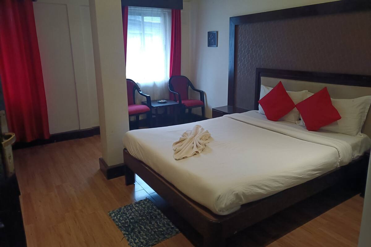 A hotel room with a bed, a small table with chairs, and a window with red curtains.