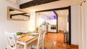 A dining area with a wooden table and white chairs, a kitchen in the background, and a trash bin to the side. - Cosy old Nice Appartement (Nice)