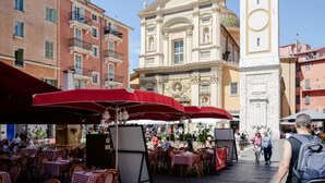 An outdoor café with tables and red umbrellas, set against a backdrop of historic buildings and a prominent church. - Cosy old Nice Appartement (Nice)
