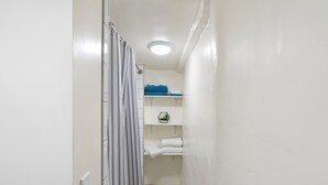 Un baño estrecho con paredes blancas, una cortina de ducha, un perchero y un estante pequeño con toallas.