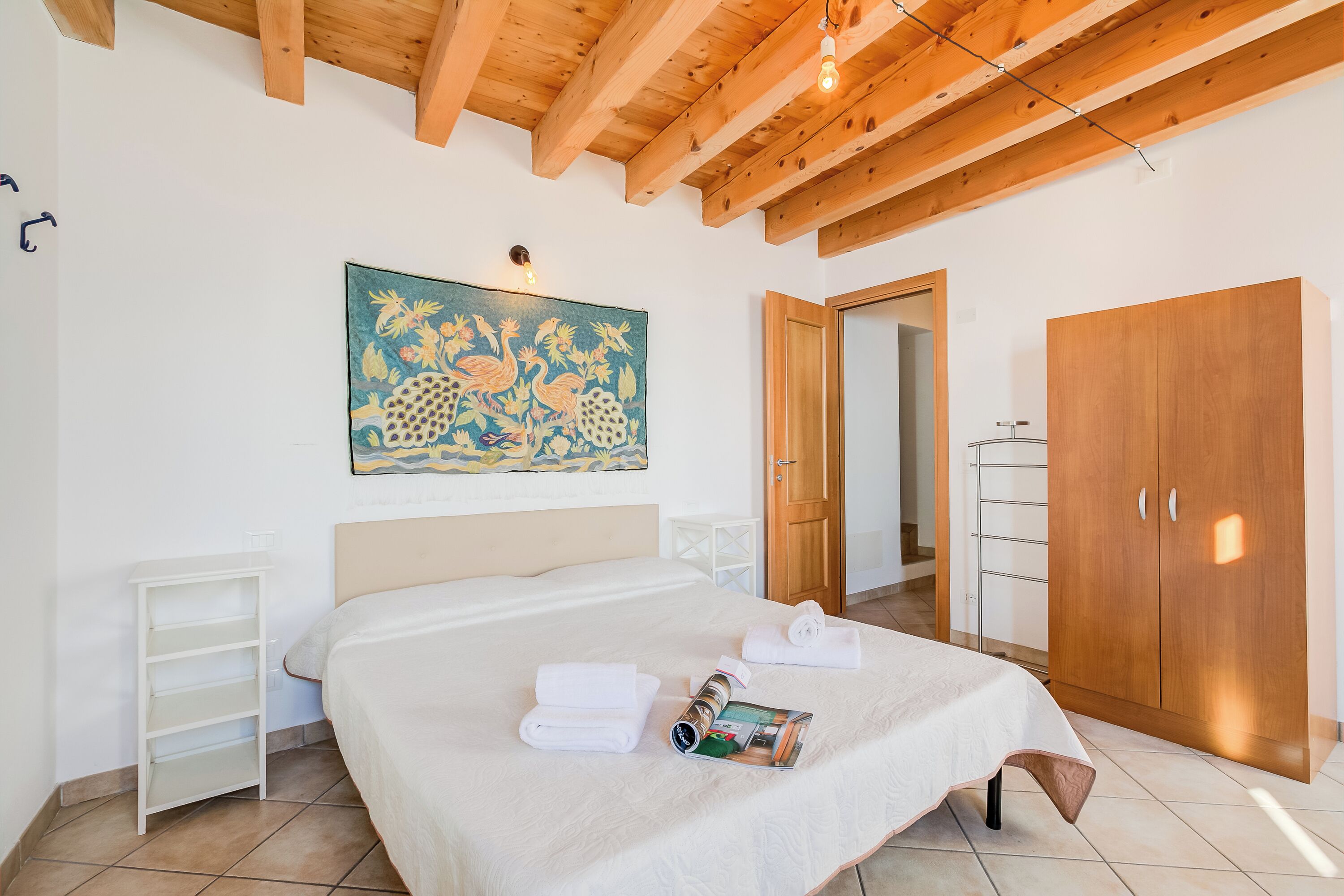 A bedroom with a bed, a wooden wardrobe, a painting on the wall, and a white bedside table.